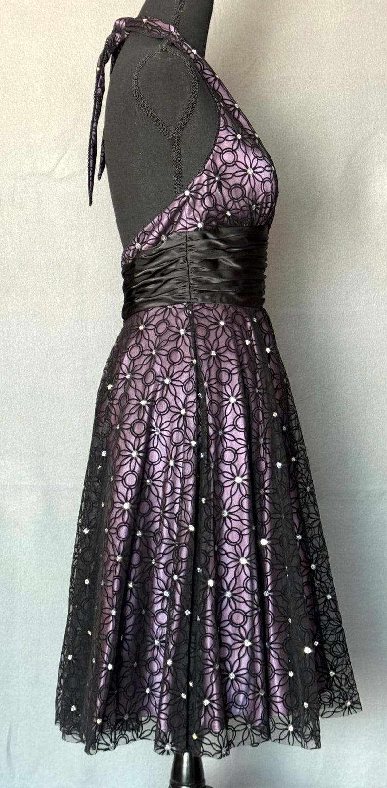 May include: A formal halter dress with a black floral lace overlay over a lavender lining. The dress features a black satin waistband and is adorned with small, sparkling embellishments.