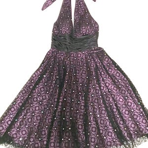 May include: A halter-neck dress in a lilac hue, overlaid with a black floral lace pattern. The dress features a black satin waistband and is adorned with small, sparkling embellishments. The skirt flares out, and the dress is suitable for special occasions.