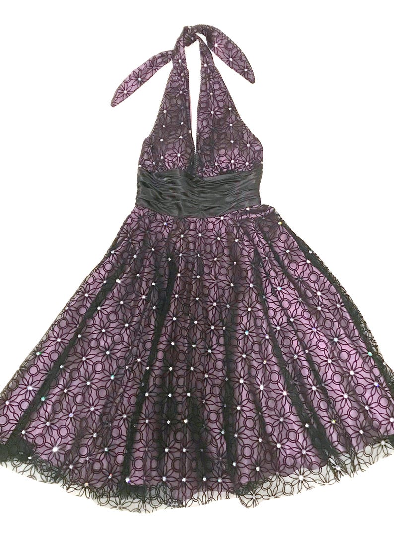 May include: A halter-neck dress in a lilac hue, overlaid with a black floral lace pattern. The dress features a black satin waistband and is adorned with small, sparkling embellishments. The skirt flares out, and the dress is suitable for special occasions.