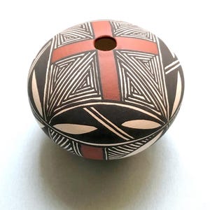 May include: A round, hand-painted ceramic vase with a cross design. The vase features a terracotta-colored cross on a black background, with white geometric patterns. The vase has a small opening at the top and is likely used for decorative purposes.