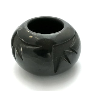 May include: A small, round, black ceramic vessel with a wide opening. The exterior features carved, stylized geometric designs. The glossy finish reflects light, highlighting the carved details. The vessel sits on a white surface.