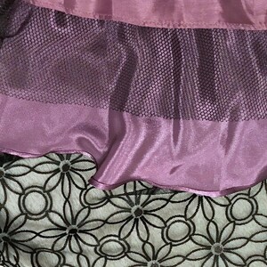 May include: Close-up of layered fabric in shades of purple and black. The top layer is a solid, shiny purple fabric. Below it is a layer of black mesh, and at the bottom is a layer of white fabric with a black floral pattern.