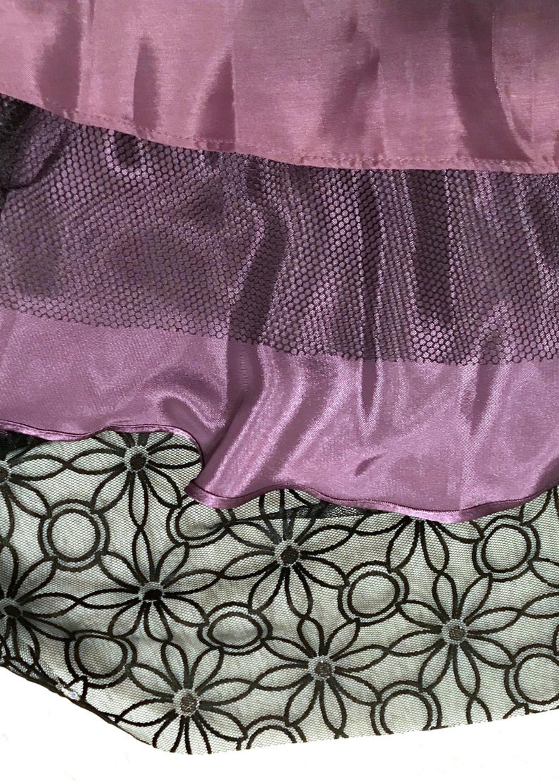 May include: Close-up of layered fabric in shades of purple and black. The top layer is a solid, shiny purple fabric. Below it is a layer of black mesh, and at the bottom is a layer of white fabric with a black floral pattern.