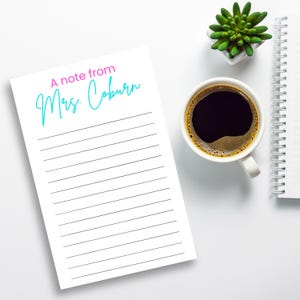 May include: A white notepad with lined pages and the text "A note from Mrs. Coburn" in pink and teal. A cup of coffee, a small succulent, and a spiral notebook are also on the white surface.
