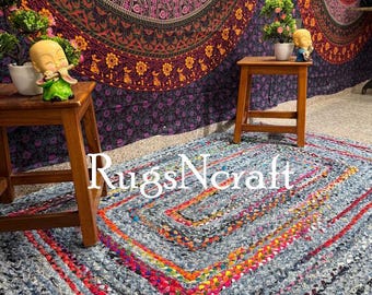 May include: A rectangular, braided rug with a denim blue base and vibrant red, orange, and yellow accents. The rug features a geometric pattern and the text "RugsNcraft". Two small wooden stools and a tapestry are in the background.