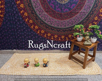 May include: A beige and white patterned rug with three small, gold-colored figurines on top. The rug is in front of a purple and gold mandala tapestry with a wooden table and potted plants.