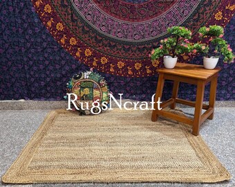 May include: A natural jute rug with a woven border in a light brown colour. The rug is laid out on a floor in front of a wall with a colourful mandala tapestry and a small wooden stool with two potted plants.