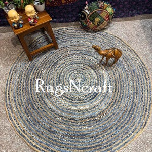 Może przedstawiać: Okrągły dywan z juty w kolorze niebieskim i naturalnym. Dywan ma koncentryczny wzór w postaci kół z jasnoniebieskim środkiem i ciemnoniebieskim zewnętrznym brzegiem. Dywan jest wykonany z naturalnych włókien juty i ma rustykalny, ręcznie tkany wygląd. Tekst "RugsNcraft" jest widoczny w środku dywanu.