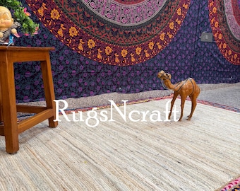 May include: A natural jute rug with a colorful woven border. The rug is laid out on a floor with a decorative tapestry hanging on the wall behind it. A small wooden stool and a camel figurine are also visible in the image.