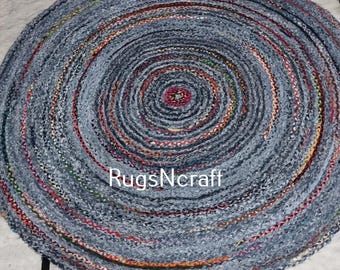 May include: A round, blue and white braided rug made from recycled denim. The rug has a circular pattern with a centre circle of red, pink, and orange. The rug is labelled "RugsNcraft".
