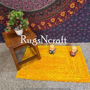 Puede incluir: Una alfombra trenzada amarilla con textura tejida. Dos pequeñas figuritas decorativas se colocan sobre la alfombra. La alfombra está delante de un tapiz morado oscuro con un patrón floral.