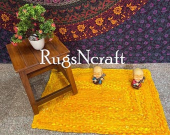 May include: A yellow braided rug with a woven texture. Two small decorative figurines are placed on the rug. The rug is in front of a dark purple tapestry with a floral pattern.