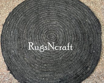Puede incluir: Una alfombra redonda de yute negro con un diseño trenzado. La alfombra tiene una forma circular y está hecha de fibras naturales. El texto "RugsNcraft" es visible en el centro de la alfombra.