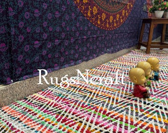 May include: A colorful, handwoven rug with a chevron pattern. The rug is made of recycled materials and is perfect for adding a touch of bohemian style to any room. The rug is laid out on a floor with a tapestry hanging on the wall behind it.