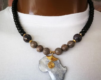 May include: A necklace featuring a pendant shaped like the continent of Africa, crafted from grey stone with a gold-toned wire spiral accent. The chain consists of smooth black beads, transitioning into speckled brown and dark grey stone beads.