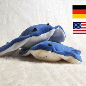 Könnte beinhalten: Zwei blaue Plüschtiere in Form von Mantarochen mit weißen Unterseiten und schwarzen Augen werden auf einer weißen, strukturierten Oberfläche gezeigt. Die Mantarochen liegen übereinander. Das Bild enthält die amerikanische und die deutsche Flagge.