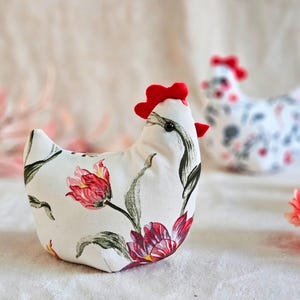 May include: Two fabric hen figurines. The primary hen is white with a floral pattern of red and pink tulips and green leaves. It has a red comb and wattle. A second hen with a floral pattern is in the background.