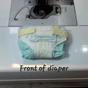May include: A white and blue baby diaper with yellow elastic bands. The diaper has the words "Pampers Baby" printed on the front. The diaper is sitting on a white surface. Front of diaper.