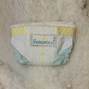 Puede incluir: Un pañal de bebé blanco con detalles amarillos y celeste. El pañal tiene la palabra "Pampers" impresa en azul claro. El pañal está diseñado para absorber líquidos y mantener al bebé seco.