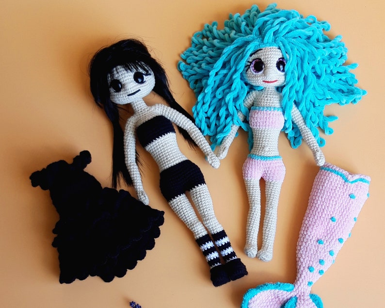 K&ouml;nnte beinhalten: Zwei handgefertigte H&auml;kelpuppen. Eine Puppe hat schwarzes Haar und tr&auml;gt ein schwarz-wei&szlig;es Outfit. Die andere Puppe hat blaues Haar und tr&auml;gt ein rosa-wei&szlig;es Outfit. Ein schwarzes Kleid und eine rosa Meerjungfrauenflosse sind ebenfalls enthalten.
