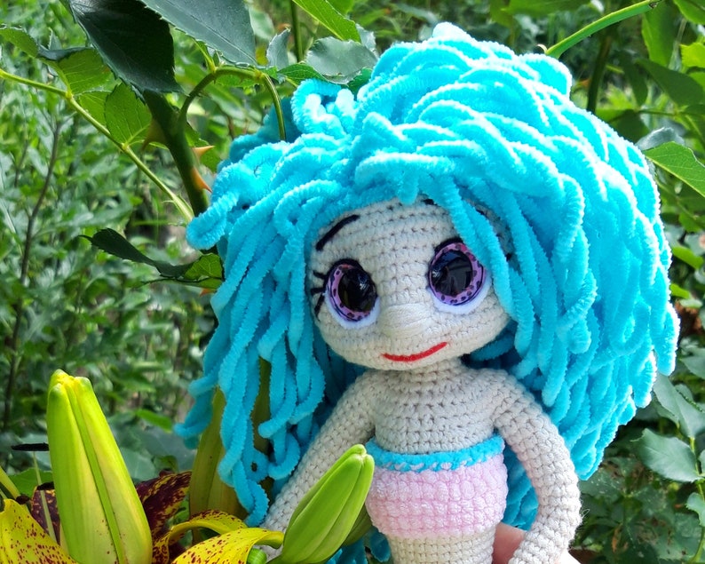 K&ouml;nnte beinhalten: Handgefertigte H&auml;kelpuppe mit hellblauem Garnhaar und gro&szlig;en, ausdrucksstarken Augen. Die Puppe hat einen hellen Hautton, einen rosa Bikini-Unterteil und befindet sich vor einem Hintergrund aus gr&uuml;nem Laub und gelben Blumen.