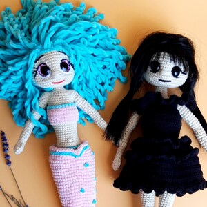 K&ouml;nnte beinhalten: Zwei handgefertigte H&auml;kelpuppen. Eine Puppe ist eine Meerjungfrau mit hellblauem Garnhaar, einem rosa Oberteil und Schwanz. Die andere Puppe hat schwarzes Haar und ein schwarzes Kleid. Beide Puppen haben gro&szlig;e Augen und sind aus beigem Garn gefertigt.