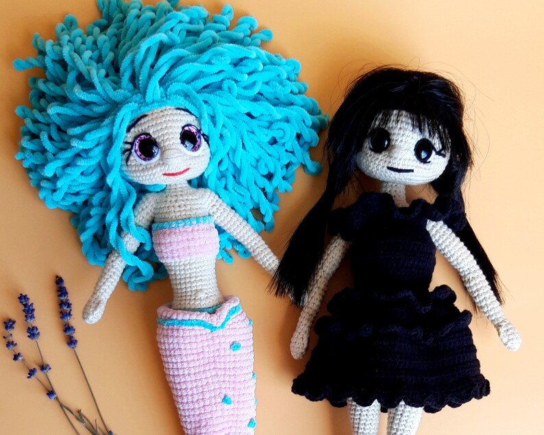 K&ouml;nnte beinhalten: Zwei handgefertigte H&auml;kelpuppen. Eine Puppe ist eine Meerjungfrau mit hellblauem Garnhaar, einem rosa Oberteil und Schwanz. Die andere Puppe hat schwarzes Haar und ein schwarzes Kleid. Beide Puppen haben gro&szlig;e Augen und sind aus beigem Garn gefertigt.