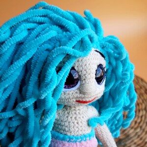 K&ouml;nnte beinhalten: Handgefertigte H&auml;kelpuppe mit leuchtend blauem, flauschigem Haar. Die Puppe hat gro&szlig;e, ausdrucksstarke Augen mit lila Akzenten, einen kleinen roten Mund und ein helles Gesicht. Der K&ouml;rper der Puppe ist cremefarben, mit einem rosa und blauen Rock.