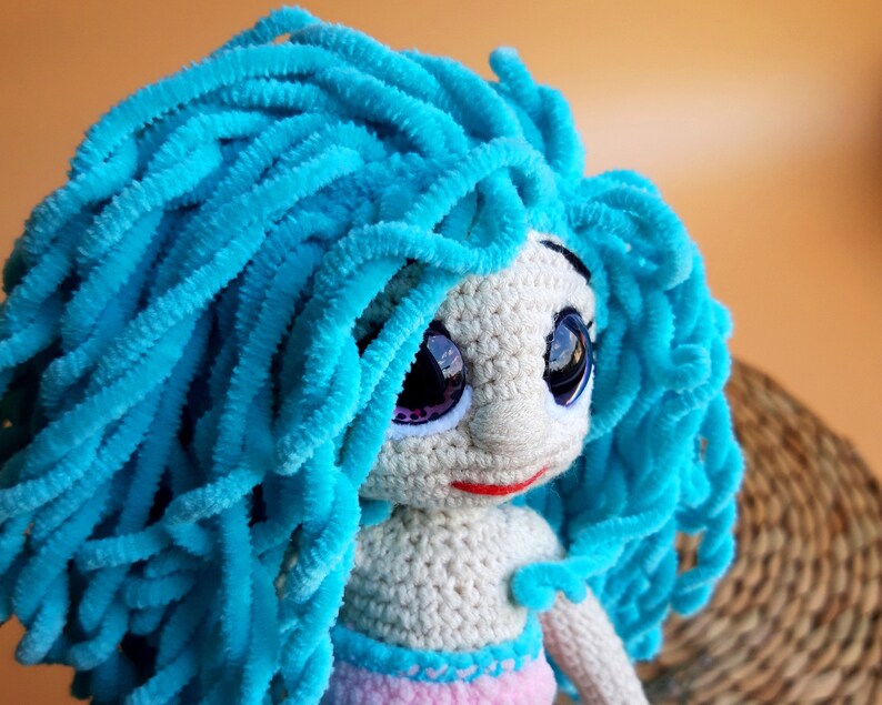 K&ouml;nnte beinhalten: Handgefertigte H&auml;kelpuppe mit leuchtend blauem, flauschigem Haar. Die Puppe hat gro&szlig;e, ausdrucksstarke Augen mit lila Akzenten, einen kleinen roten Mund und ein helles Gesicht. Der K&ouml;rper der Puppe ist cremefarben, mit einem rosa und blauen Rock.