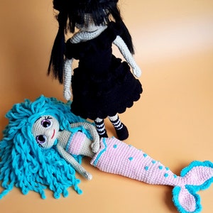 K&ouml;nnte beinhalten: Zwei handgefertigte H&auml;kelpuppen. Eine Puppe ist eine Meerjungfrau mit blauen Haaren, einem rosa Oberteil und einem rosa Schwanz mit blauen Akzenten. Die andere Puppe tr&auml;gt ein schwarzes Kleid mit schwarzen Haaren und gestreiften Socken.