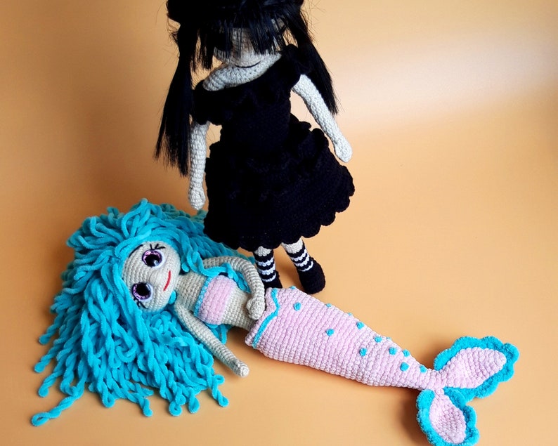 K&ouml;nnte beinhalten: Zwei handgefertigte H&auml;kelpuppen. Eine Puppe ist eine Meerjungfrau mit blauen Haaren, einem rosa Oberteil und einem rosa Schwanz mit blauen Akzenten. Die andere Puppe tr&auml;gt ein schwarzes Kleid mit schwarzen Haaren und gestreiften Socken.