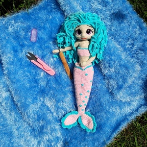 K&ouml;nnte beinhalten: Eine handgefertigte H&auml;kelpuppe in Form einer Meerjungfrau mit t&uuml;rkisfarbenem Haar und einem rosa Oberteil und Schwanz. Die Puppe liegt auf einer blauen, flauschigen Oberfl&auml;che, mit einer rosa Schere und einem H&auml;kelhaken aus Holz. Die Meerjungfrau hat gro&szlig;e, ausdrucksstarke Augen.