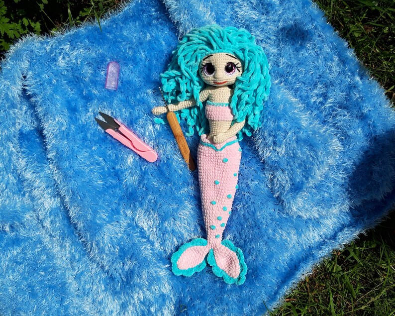 K&ouml;nnte beinhalten: Eine handgefertigte H&auml;kelpuppe in Form einer Meerjungfrau mit t&uuml;rkisfarbenem Haar und einem rosa Oberteil und Schwanz. Die Puppe liegt auf einer blauen, flauschigen Oberfl&auml;che, mit einer rosa Schere und einem H&auml;kelhaken aus Holz. Die Meerjungfrau hat gro&szlig;e, ausdrucksstarke Augen.