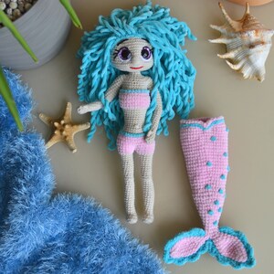 K&ouml;nnte beinhalten: Handgefertigte H&auml;kelpuppe einer Meerjungfrau mit blauen Garnhaaren, die ein rosa Bikini-Oberteil und Shorts tr&auml;gt. Ein separater rosa Meerjungfrauenschwanz mit blauen Akzenten ist ebenfalls enthalten. Die Puppe ist etwa 20 cm gro&szlig;.