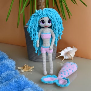 K&ouml;nnte beinhalten: Handgefertigte H&auml;kelpuppe einer Meerjungfrau mit blauen Haaren, die ein rosa Bikini-Oberteil und Shorts tr&auml;gt. Die Puppe hat einen passenden rosa und blauen Schwanz. Ein Seestern und eine Muschel liegen neben der Puppe.