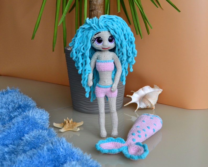 K&ouml;nnte beinhalten: Handgefertigte H&auml;kelpuppe einer Meerjungfrau mit blauen Haaren, die ein rosa Bikini-Oberteil und Shorts tr&auml;gt. Die Puppe hat einen passenden rosa und blauen Schwanz. Ein Seestern und eine Muschel liegen neben der Puppe.