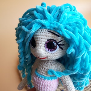 K&ouml;nnte beinhalten: Handgefertigte H&auml;kelpuppe in Form einer Meerjungfrau mit hellblauem, flauschigem Haar. Die Puppe hat gro&szlig;e, glitzernde Augen und ein rosa-blaues Outfit. Die Haut der Puppe ist hellbeige und sie hat einen kleinen roten Mund.