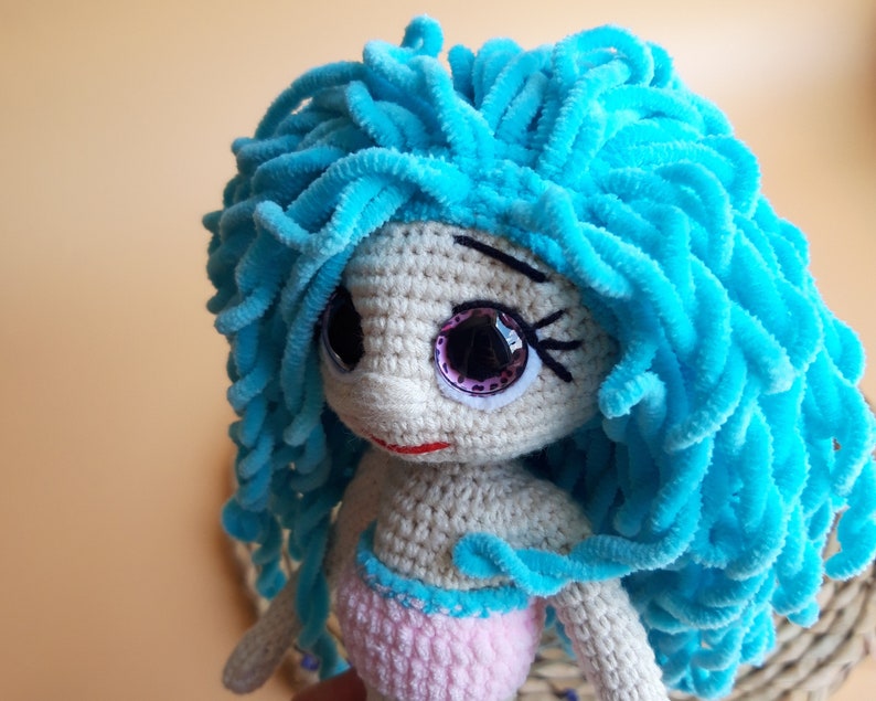 K&ouml;nnte beinhalten: Handgefertigte H&auml;kelpuppe in Form einer Meerjungfrau mit hellblauem, flauschigem Haar. Die Puppe hat gro&szlig;e, glitzernde Augen und ein rosa-blaues Outfit. Die Haut der Puppe ist hellbeige und sie hat einen kleinen roten Mund.