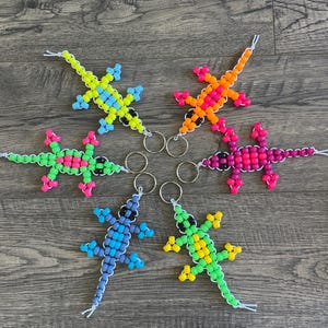 May include: Six beaded lizard keychains in vibrant colors: yellow, orange, pink, green, purple, and blue. Each lizard has a contrasting color for its feet and is attached to a silver keyring. The keychains are arranged on a wooden surface.
