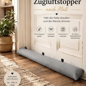 Könnte beinhalten: Ein langer, zylindrischer Zugluftstopper aus grauem Stoff, der vor einer weißen Tür liegt. Er ist mit kleinen schwarzen Stoffschlaufen an der Oberkante versehen. Der Text auf dem Bild lautet 'Zugluftstopper nach Maß' und nennt Längen von 40 bis 200 cm.