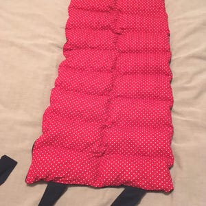 May include: A red polka dot heating pad with black straps. The rectangular pad is made of quilted fabric and is designed to be worn around the neck or shoulders. The straps are attached to the top and bottom of the pad.
