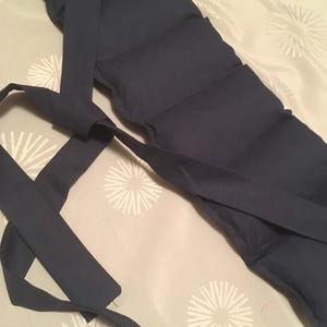 May include: A navy blue, segmented wrap with long ties attached. The wrap is made of a soft fabric and is designed to be tied around the body. The background is a light beige fabric with a white floral pattern.