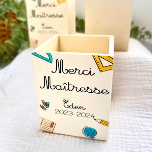 Peut inclure: Un porte-crayons en bois avec un fond blanc et des illustrations colorées sur le thème de l'école. Le texte "Merci Maitresse Eden 2023-2024" est imprimé sur le devant.