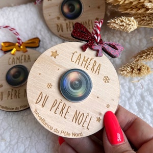 Peut inclure: Une décoration de Noël en bois en forme d'appareil photo avec le texte "Camera du Père Noël Surveillance du Pôle Nord". La décoration est ornée d'un ruban rouge et blanc et d'un nœud rouge pailleté.