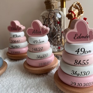 Peut inclure: Trois anneaux empilables en bois rose et blanc avec des sommets en forme de cœur. Les anneaux sont décorés de noms, de mesures en centimètres et de dates. Les noms sont "Chloe", "Alice" et "Edmée".