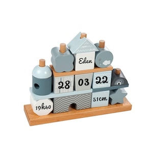 Peut inclure: Un jouet d'empilage en bois bleu et gris avec le nom "Eden", la date "28-03-22" et l'heure "19h40" imprimés sur les blocs. Le jouet comprend également une étoile, un arc-en-ciel et un nuage.