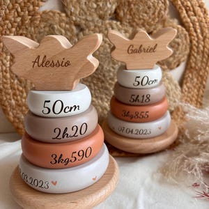 Peut inclure: Deux jouets à empiler en bois avec des dessus en forme d'ailes d'ange. L'un est étiqueté "Alessio" et l'autre "Gabriel". Chacun a des anneaux empilés avec des mesures et des dates, dont 50cm, 2h20, 3kg590 et 03.2023. Les jouets sont sur une base en bois.