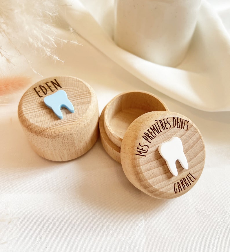 May include: Two wooden boxes with lids for storing baby teeth. The box on the left has a blue tooth icon and the name "EDEN" engraved on the lid. The box on the right has a white tooth icon and the text "MES PREMIERES DENTS GABRIEL" engraved on the lid.