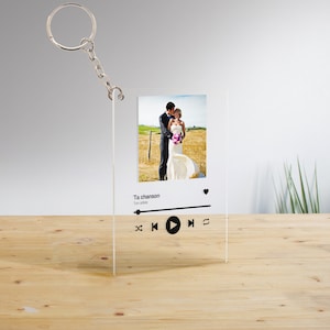 Peut inclure: Un porte-clés en acrylique transparent avec une photo d'un couple dans un champ. Le porte-clés a un design de lecteur de musique avec le texte "Ta chanson" et les mots "Ton artiste" en dessous. Le lecteur de musique a des boutons de lecture, de pause et de saut.