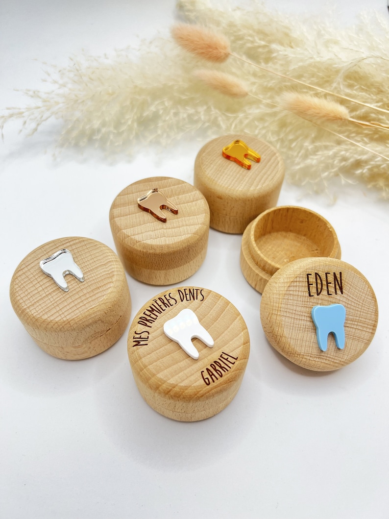 May include: Four wooden boxes with lids for storing baby teeth. Each box has a different coloured tooth charm and is personalised with a name. The boxes are on a white surface with dried pampas grass.